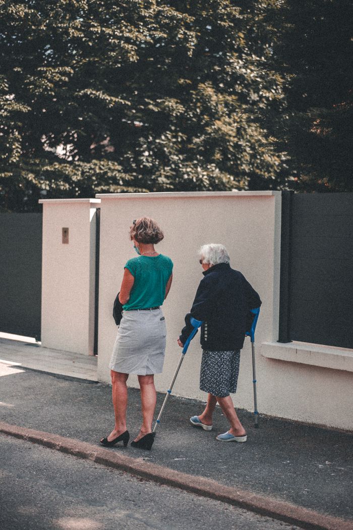 heros-img Senior woman with crutches walking alongside a companion on a sunny day in Lyon, France.