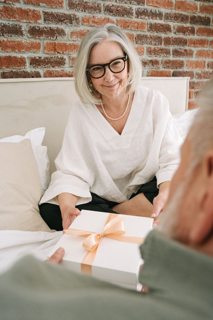 Elderly couple enjoying a moment of gift-giving in a cozy bedroom setting.