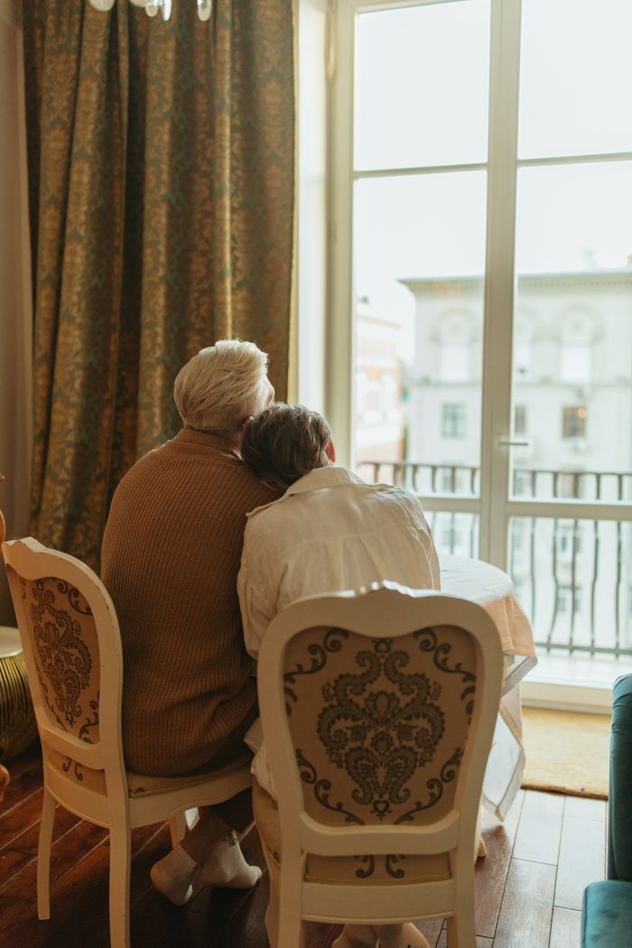 Elderly couple sitting closely together looking out a window, symbolizing love and companionship.